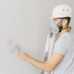 A construction worker wearing a gas mask and hard hat applies plaster to a wall indoors.